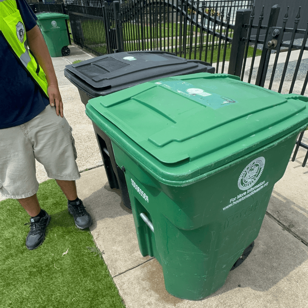 Man in a safety vest standing next to green and black outdoor wheelie bins, illustrating professional trash can cleaning services by Space City Sanitizers.