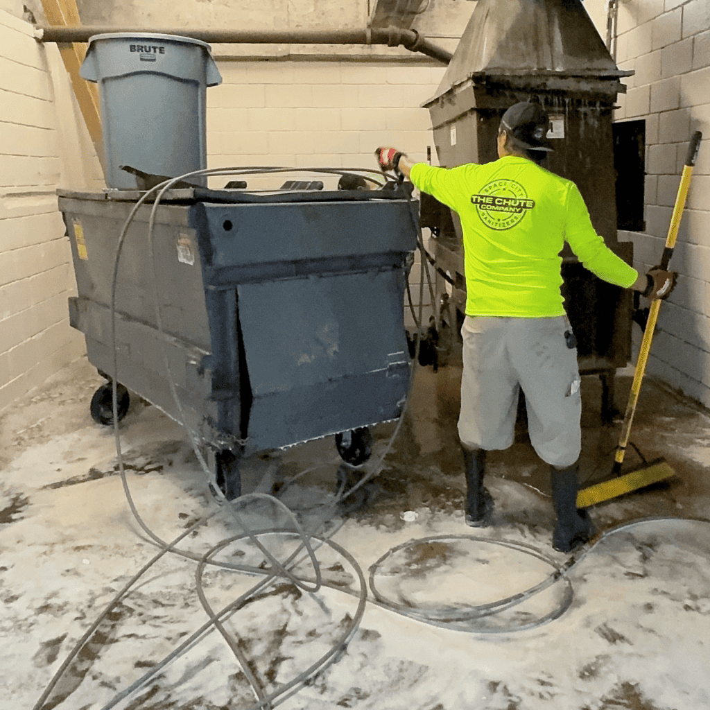 Worker cleaning a large commercial trash bin with a broom and hose in a sanitary environment, emphasizing professional trash can cleaning services.
