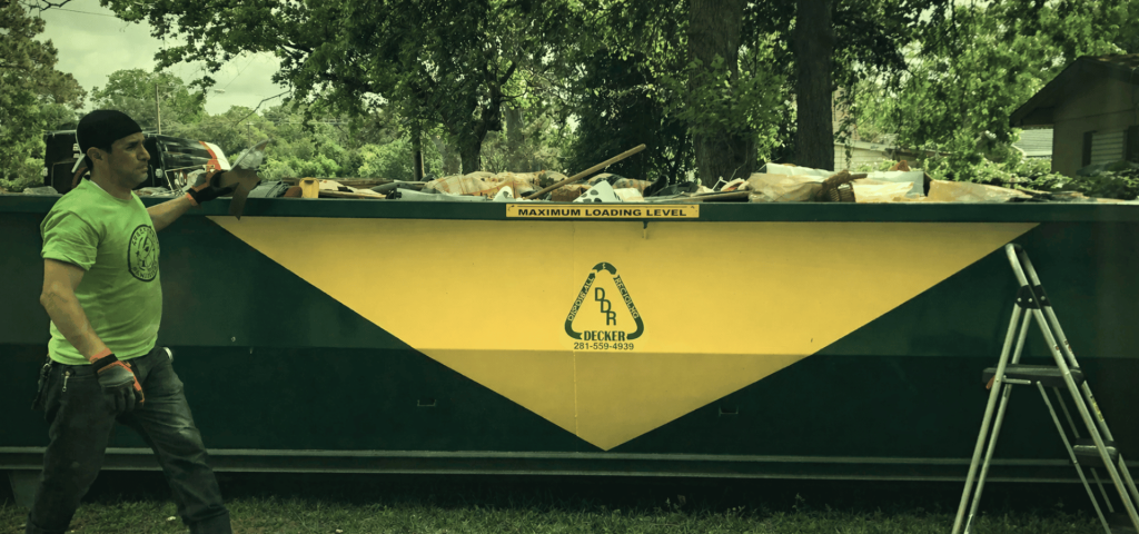 Man in green shirt and gloves standing next to a large dumpster filled with debris, illustrating post-eviction cleanup services by Space City Sanitizers.